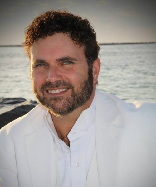 A man wearing a white suit smiles by the sea at sunset.