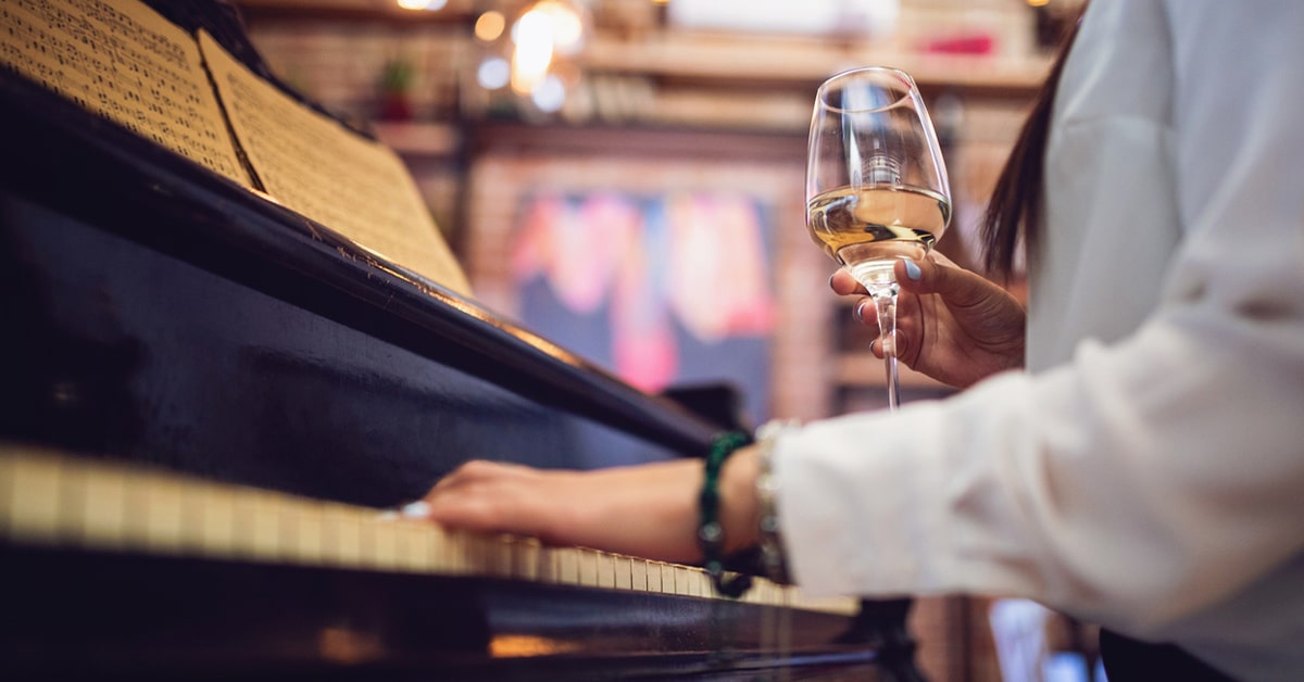 A woman playing piano with a glass of white wine in hand.