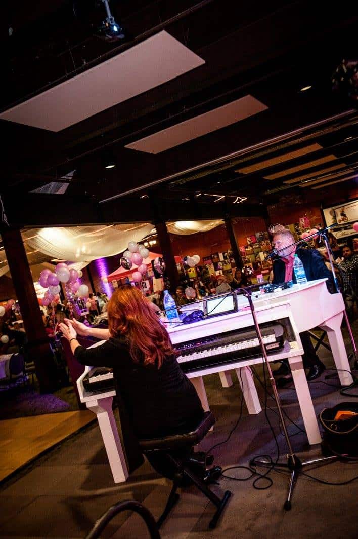 A woman photographing a man playing piano at an event with colorful balloons.