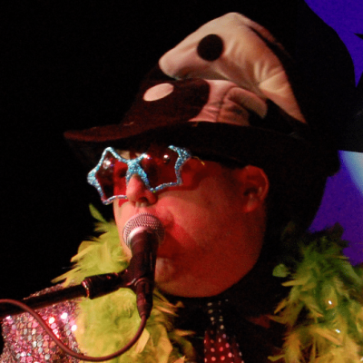 A person in colorful costume, wearing star glasses and a feather boa, singing into a microphone.