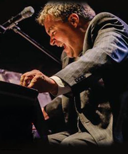 Man in a suit passionately playing piano on stage with a microphone nearby.