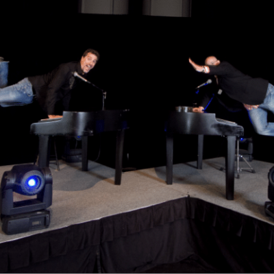 Two men in jeans and jackets humorously posing on pianos on stage with blue spotlights.