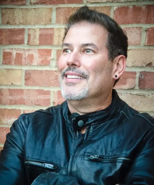 A man in a black leather jacket smiling against a brick wall background.