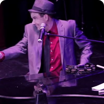 A man in a suit and hat plays the piano on stage with a microphone.